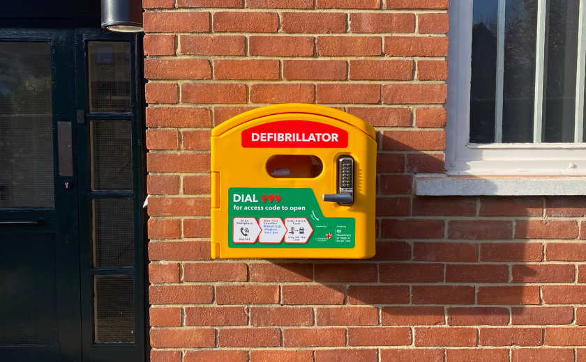 Defibrillator located in bright yellow box with green sticker indicating to dial 999 for access code.